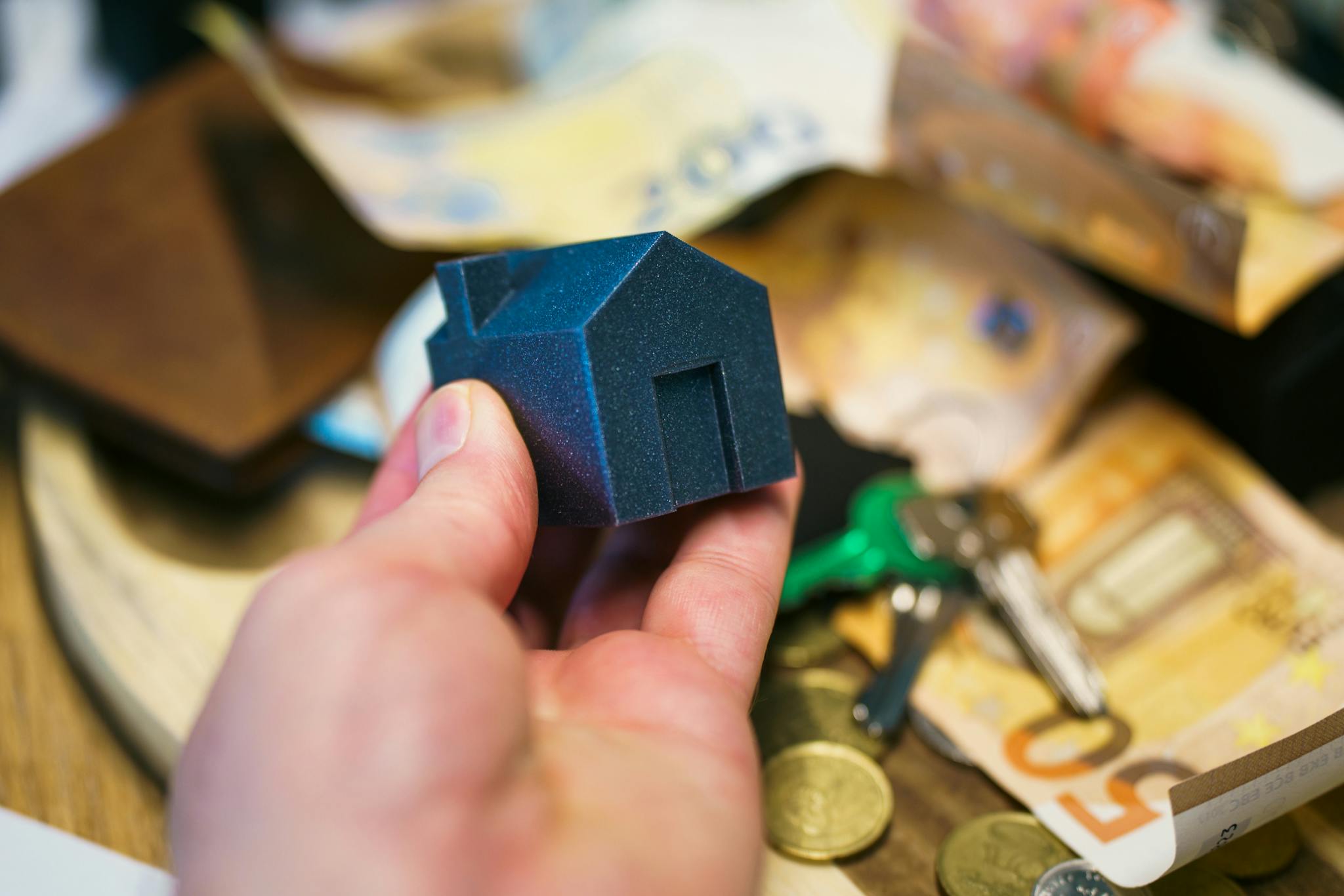 Close-up of a hand holding a small house model surrounded by euro banknotes and keys, symbolizing real estate investment.